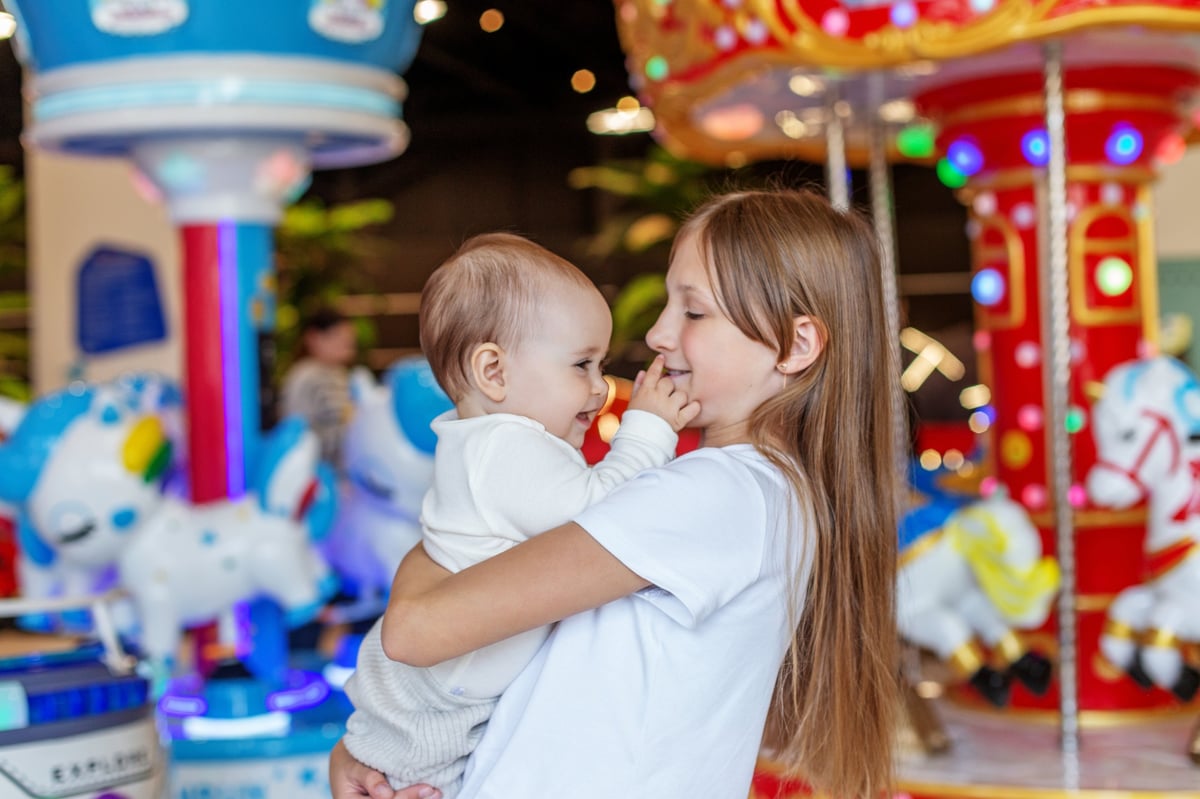 Smiling baby and sister near indoor carousel with colorful lights