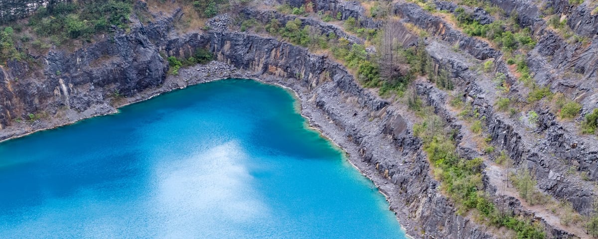 Scenic aerial landscape of an old limestone quarry featuring terraced rock walls and a deep blue water pond, surrounded by tropical forest and mountains.