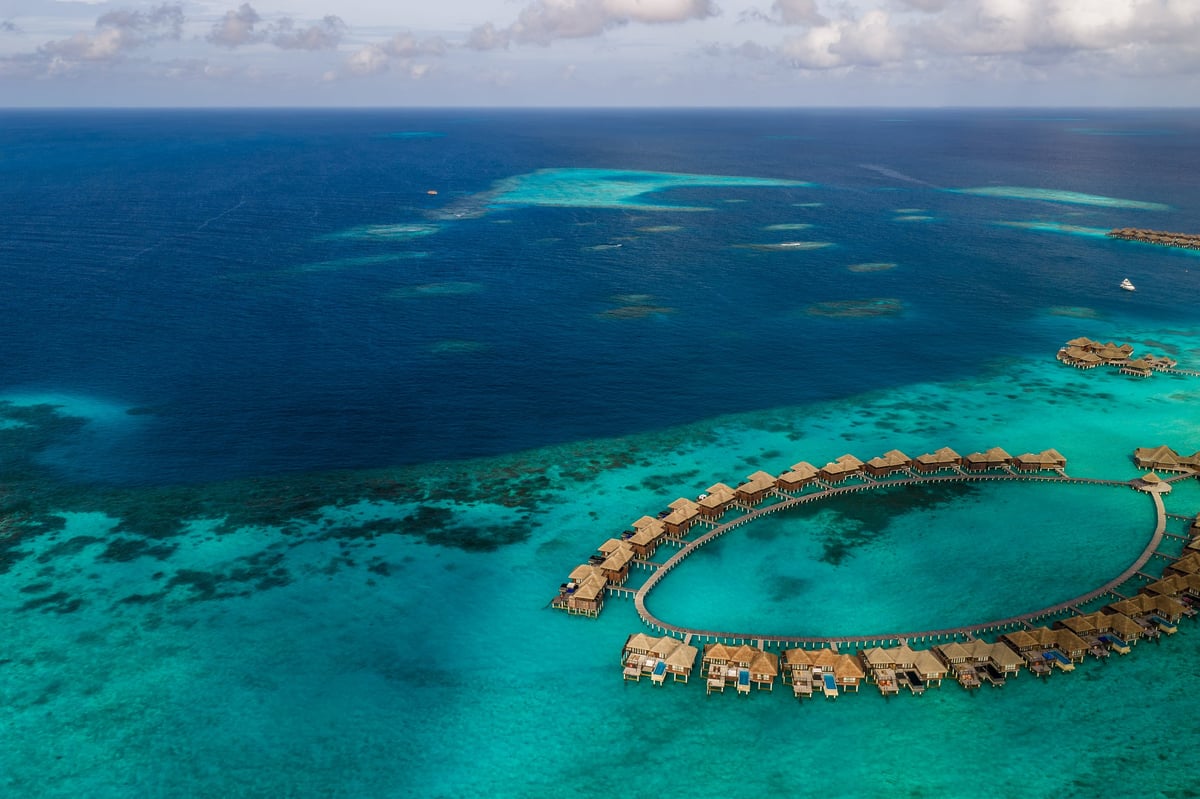 Aerial Maldives resort shows ring shaped overwater villas, circular boardwalk, thatched bungalows with decks and pools in turquoise lagoons, with darker reef patches.
