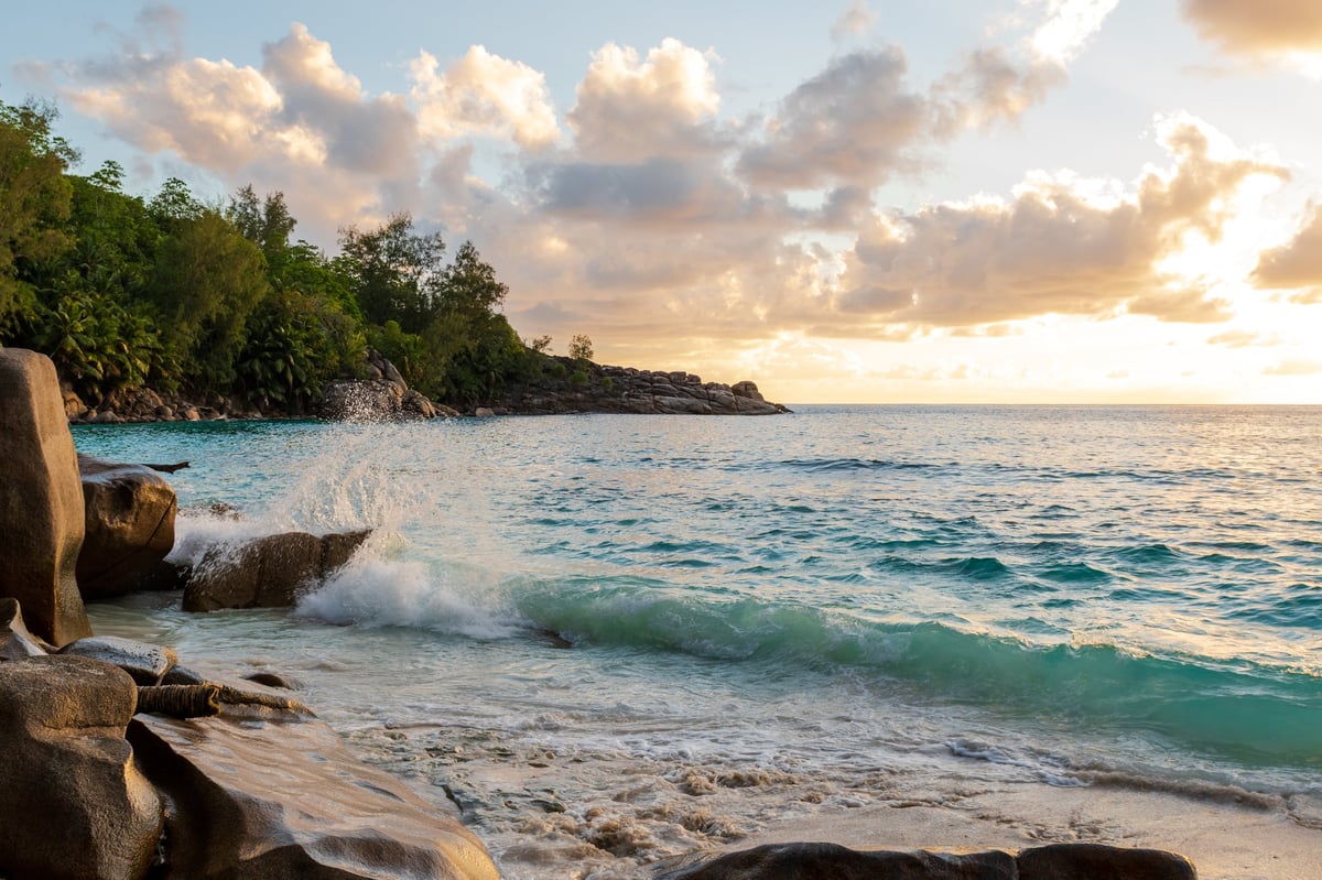 Turquoise sea surf and granite boulders at a tropical beach during golden sunset with dramatic clouds.