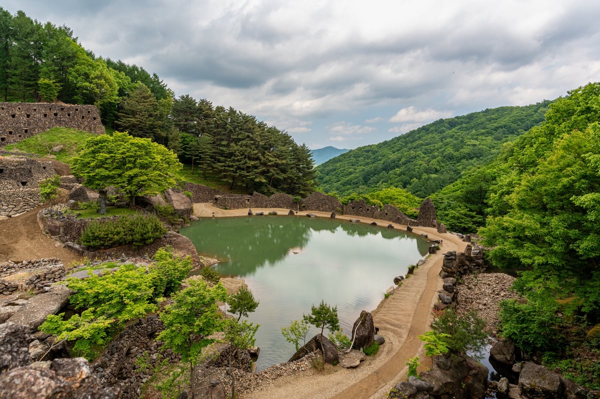A tranquil pond surrounded by stone structures, walking paths and dense green forest under a dramatic cloudy sky.