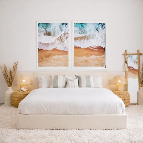 Minimalist bedroom with beige upholstered bed, white bedding, decorative pillows, woven nightstands, candles, and two coastal beach art prints above headboard
