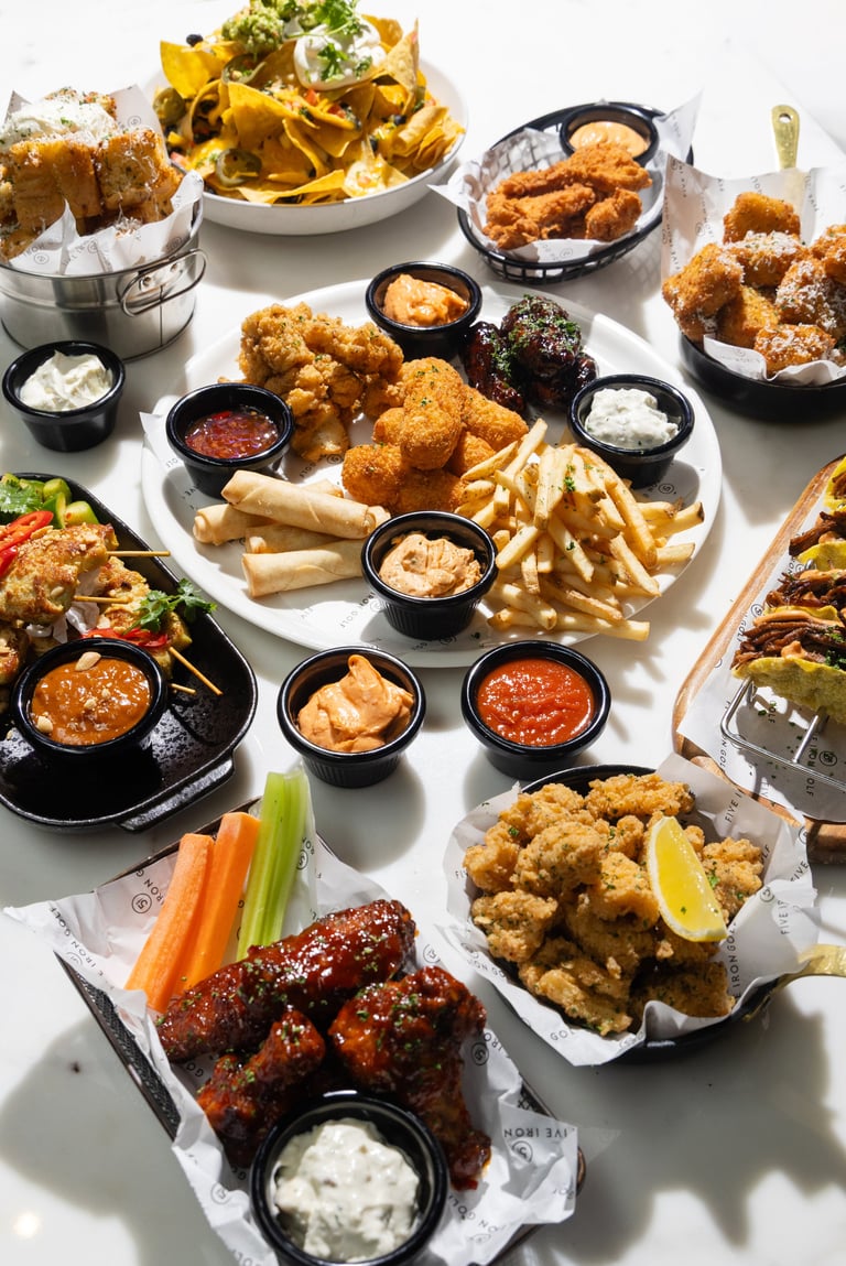 Overhead view of various fried foods including chicken wings, fried calamari, breaded shrimp, fries, and nachos with dipping sauces arranged on a white surface