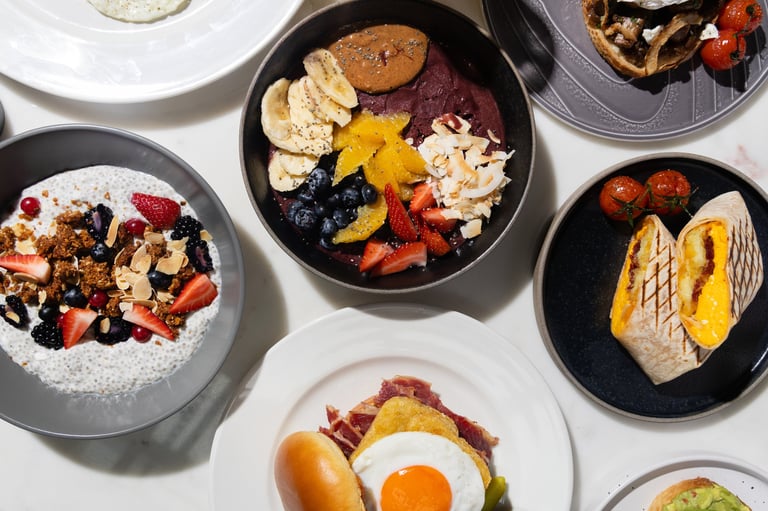 Overhead view of six bowls with assorted breakfast and brunch dishes including granola bowls, fried eggs, toast, and fresh fruits on a white surface