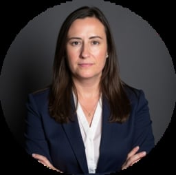 Professional headshot of a woman with long brown hair wearing a dark blue blazer and white shirt against a gray circular background