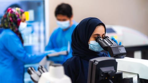 Female scientist in hijab and medical mask using microscope in laboratory with colleagues in background wearing blue coats