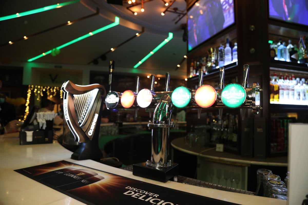 Modern bar counter with illuminated beer taps and harp display, neon green ceiling lights and TV screens in background