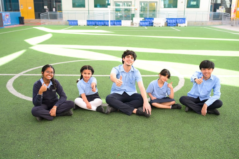 Five students sitting cross-legged on a soccer field with white markings and school building in background
