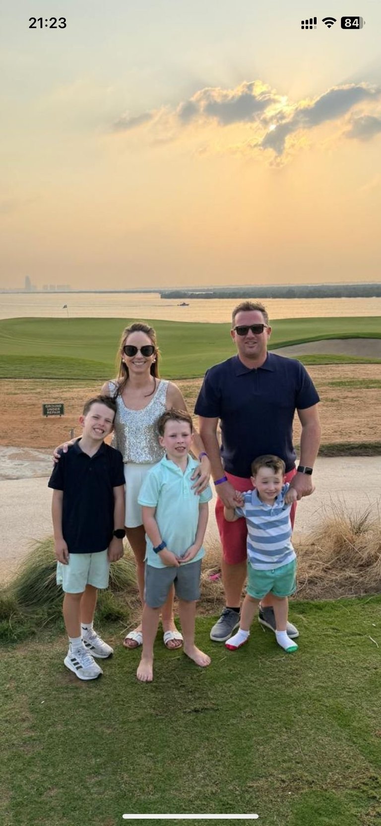 Family of five posing on grass overlooking a scenic waterside landscape with fields and sunset sky