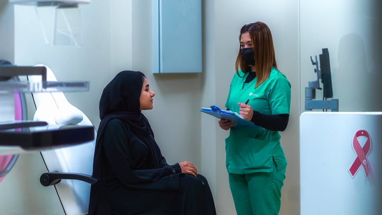 Healthcare worker in green scrubs and mask speaking with seated patient in medical office setting