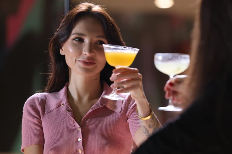 Woman in pink shirt holding up a yellow cocktail glass while toasting with another person at an indoor venue