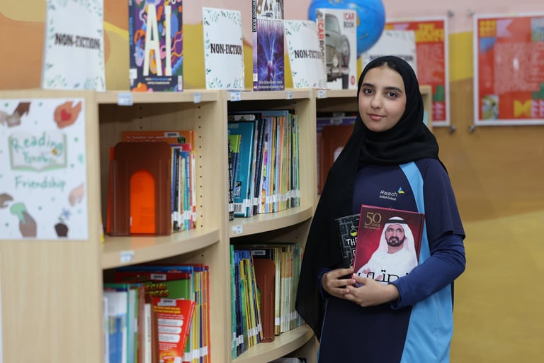 Young woman wearing a hijab holding a book while standing in front of library shelves filled with books and educational materials