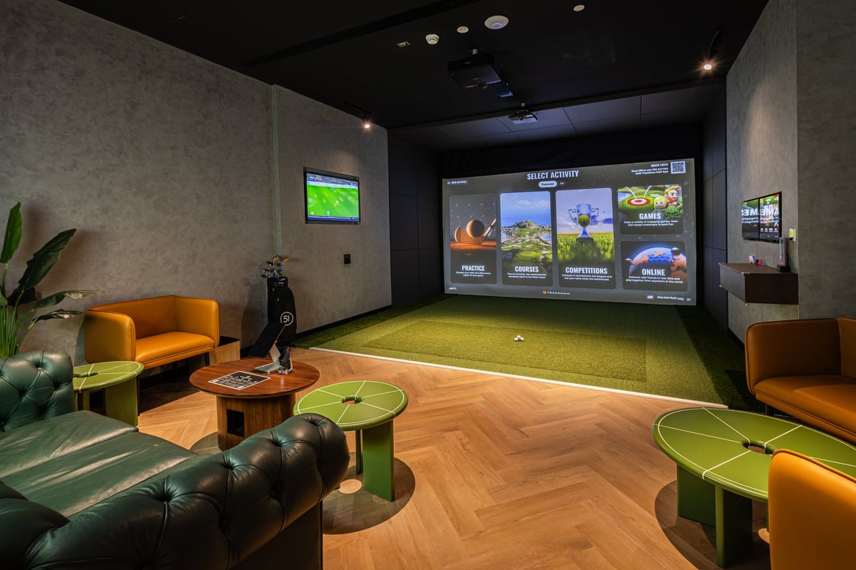 Indoor golf simulator room with a green putting area, large screen display, orange and black seating, wooden flooring, and ambient lighting