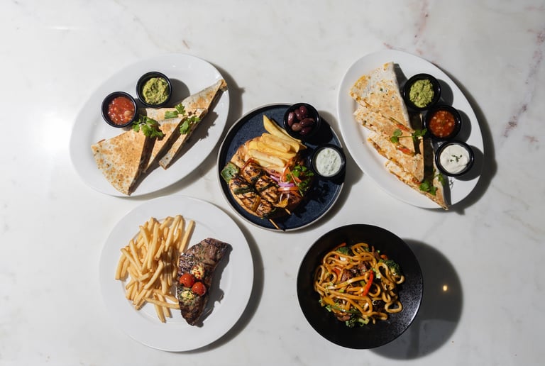 Overhead view of five plates with various Asian dishes including noodles, grilled meat, fries, and dipping sauces on a marble surface