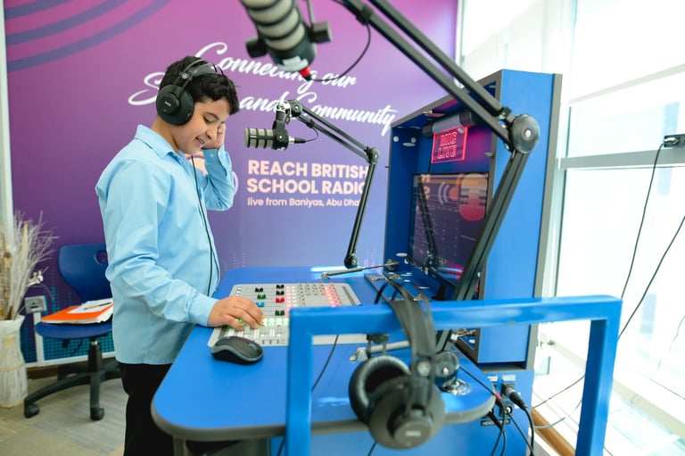 Man in light blue shirt reading at blue radio station desk with microphone and headphones visible