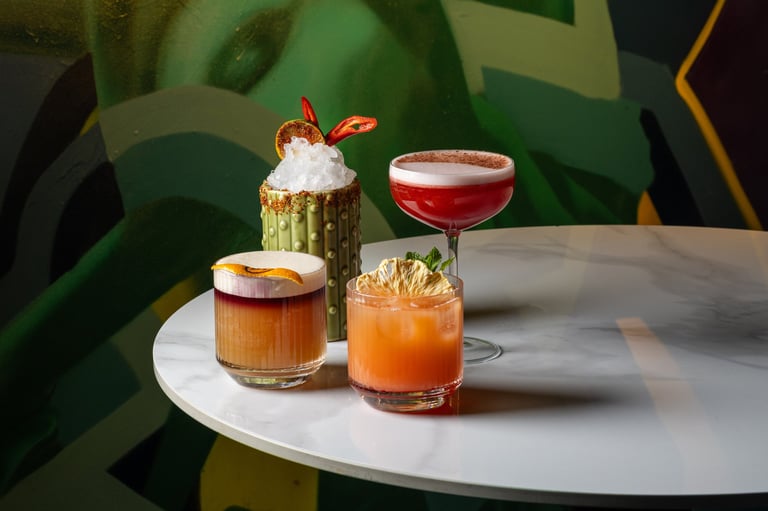 Four colorful cocktails on a white table against a green leafy backdrop, including drinks with orange, red, and amber hues with garnishes