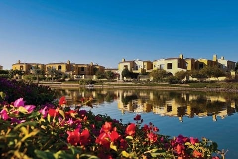 Waterfront village with terracotta buildings reflecting in calm blue water, red flowers in foreground, clear sky