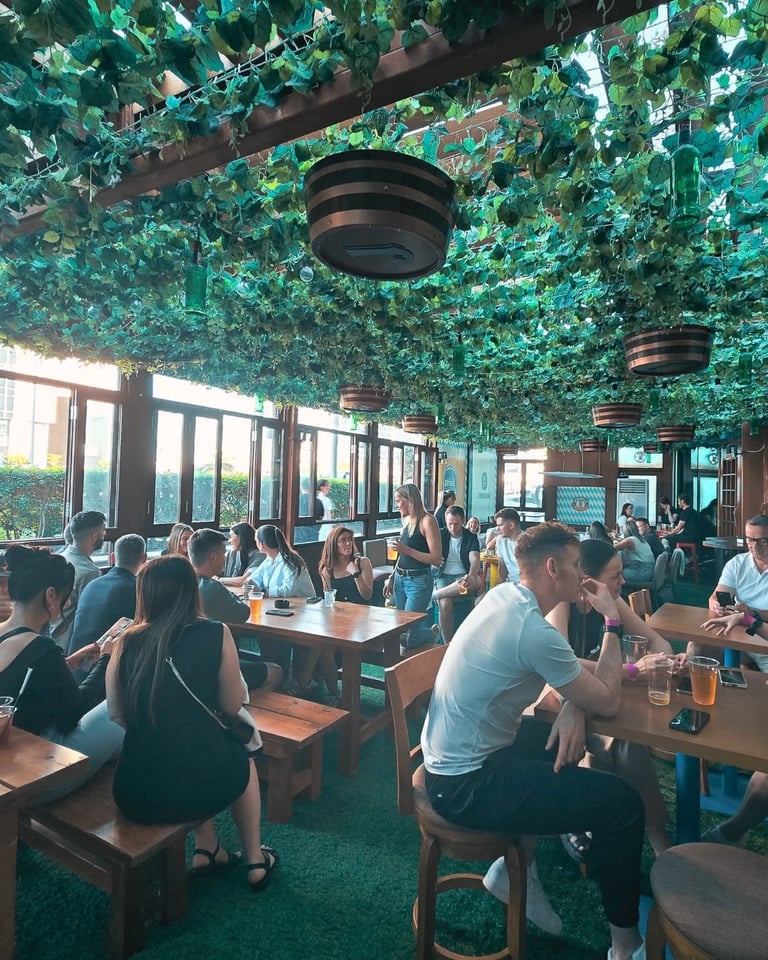 Busy restaurant with green ivy-covered ceiling, multiple diners seated at wooden tables, and natural light from large windows