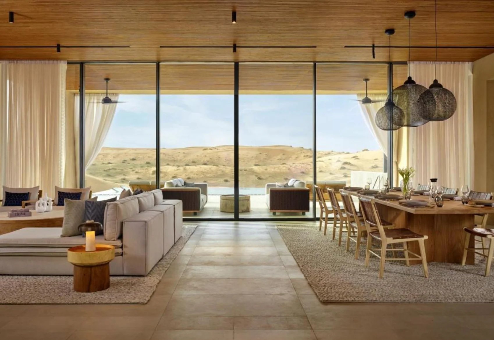Modern open-concept living space with floor-to-ceiling windows overlooking desert landscape, featuring contemporary furniture and wooden dining table