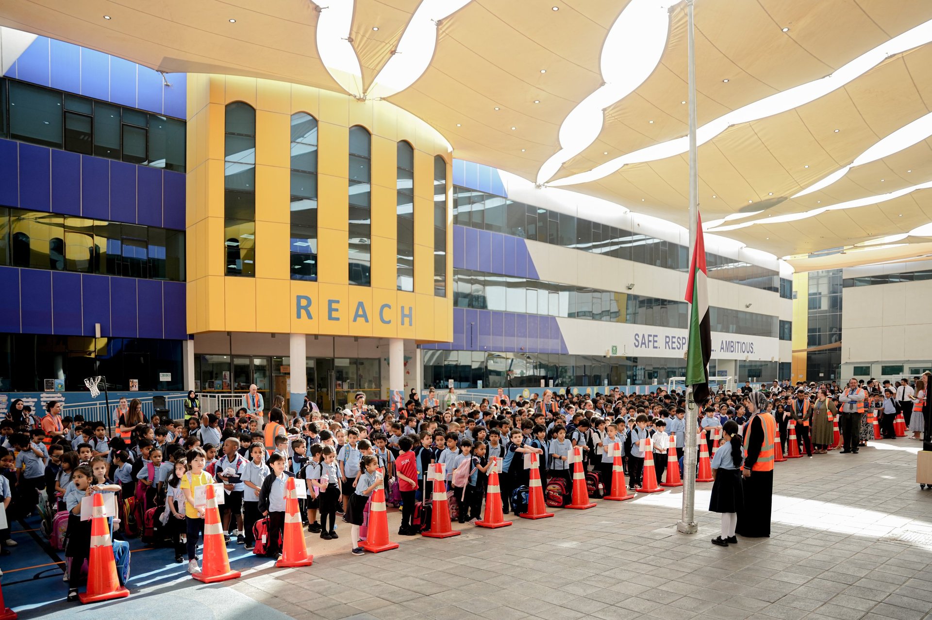Large crowd of people gathered in a modern indoor venue with yellow building and orange safety barriers — Reach British School community event