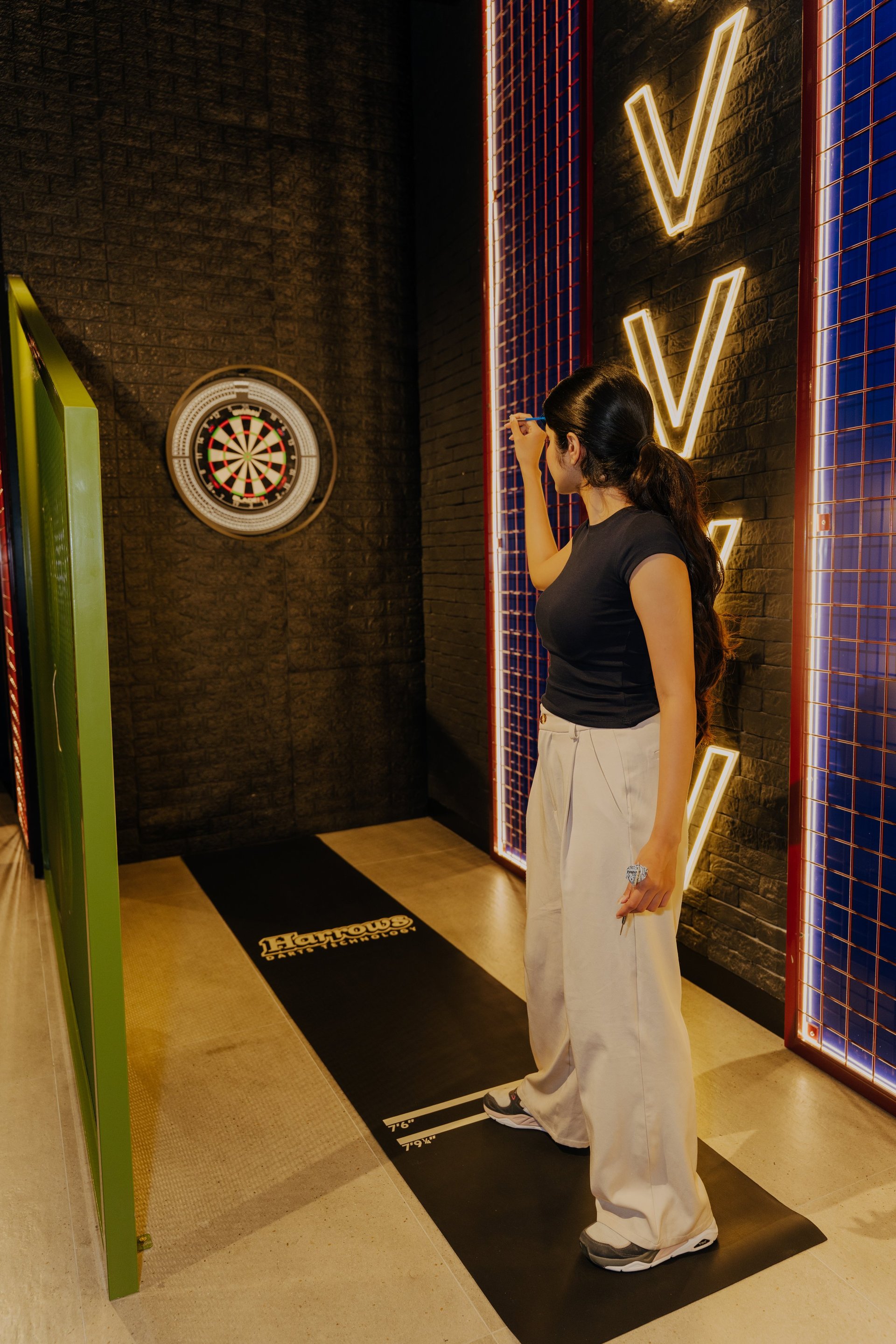 Woman playing darts in an indoor entertainment venue with neon-lit walls and a dartboard on the brick wall