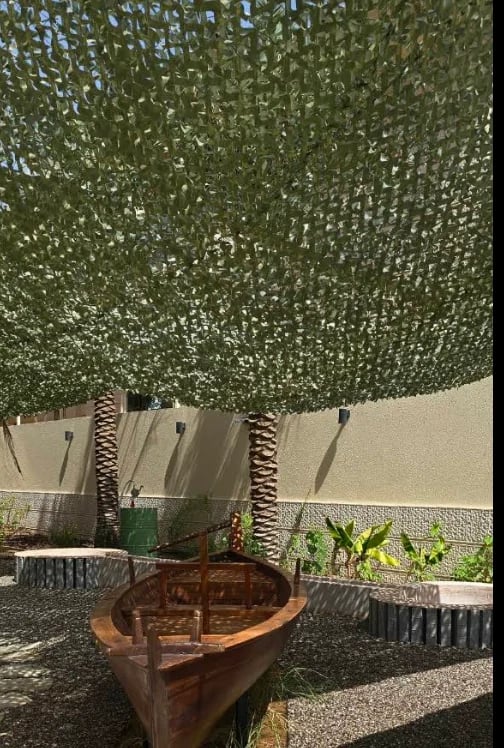 Rusty wooden rowboat displayed under green climbing ivy canopy in a courtyard with palm trees and beige walls