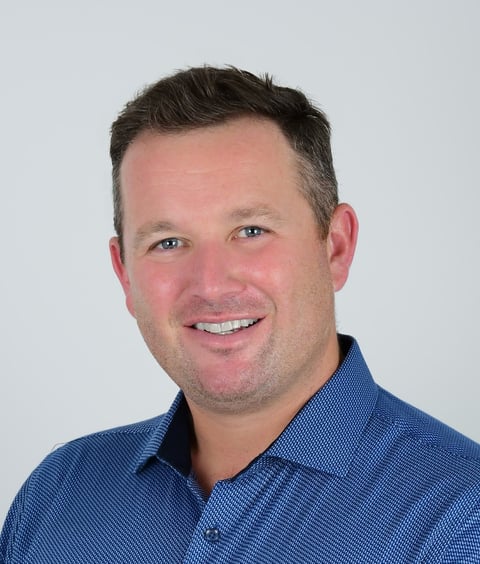 Headshot of a smiling man with dark hair wearing a blue button-up shirt against a light gray background