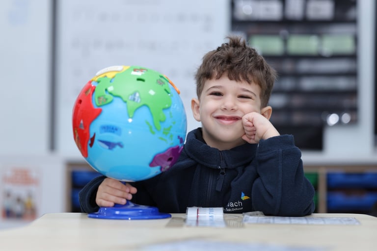 Young boy holding a colorful globe ball while smiling at a desk in a classroom setting