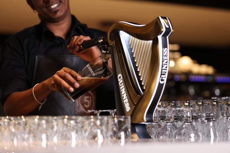 Bartender pouring a drink from a bottle behind a bar counter with glasses and a Guinness tap visible