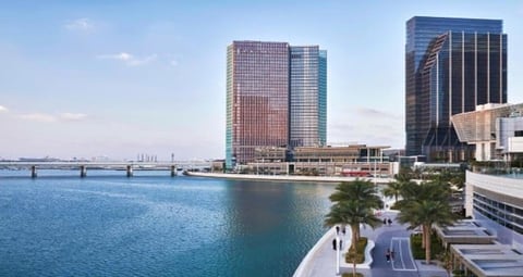 Waterfront view of modern high-rise buildings alongside a blue bay with palm trees and a promenade in the foreground